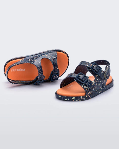 An angled front and top view of a pair of blue/orange Mini Melissa Wide Sandals with a blue base with a splatter painted print, and an orange insole.