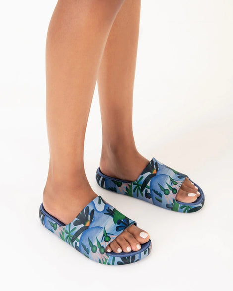 A model's legs wearing a pair of blue multicolor Melissa Cloud slides with an abstract painting design of plants, water and lagoon.