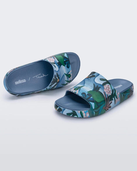 An angled front and top view of blue multicolor Melissa Cloud slides with an abstract painting design of plants, water and lagoon.