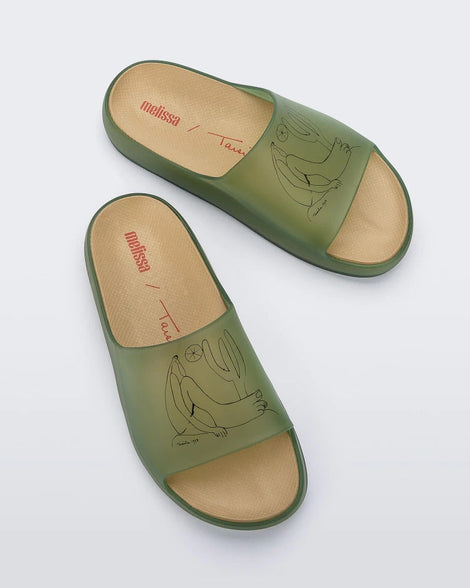 A top view of a pair of transparent green Melissa Cloud slides with a drawing of a man sitting on the ground next to a cactus and sun on the front.