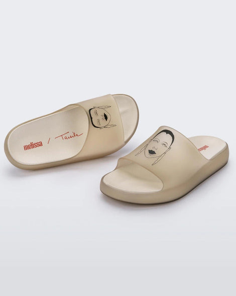 An angled top and side view of a pair of transparent beige Melissa Cloud slides with a drawing of a woman's face on the front.