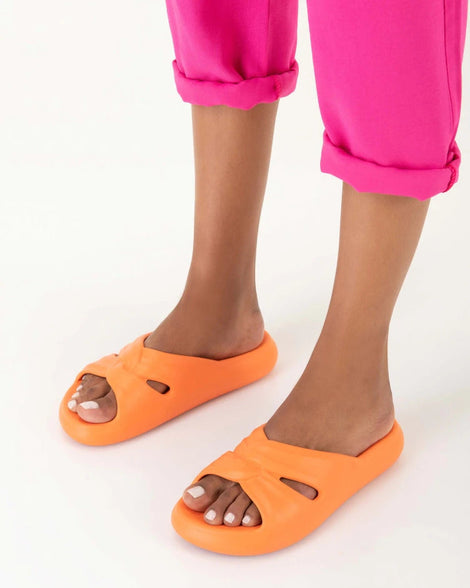 A model's legs wearing a pair of orange Melissa Free Slides with a twist front detail.