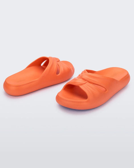 An angled back and front view of a pair of orange Melissa Free Slides with a twist front detail.