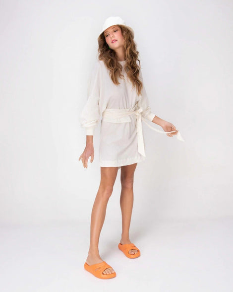 A model in a white dress posing for a picture wearing a pair of orange Melissa Free Slides with a twist front detail.