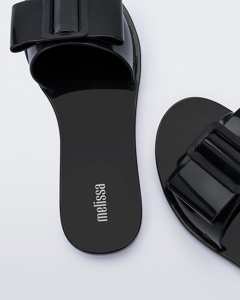 A zoomed in view of a pair of black Melissa Babe slides with a buckle like bow detail on the front strap, showing the melissa logo on the insole and an m logo on the outter strap.