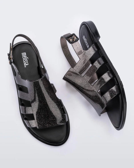 Top and side view of a pair of transparent black glitter Melissa Boemia sandals with straps conjoining in the front and a back buckle.