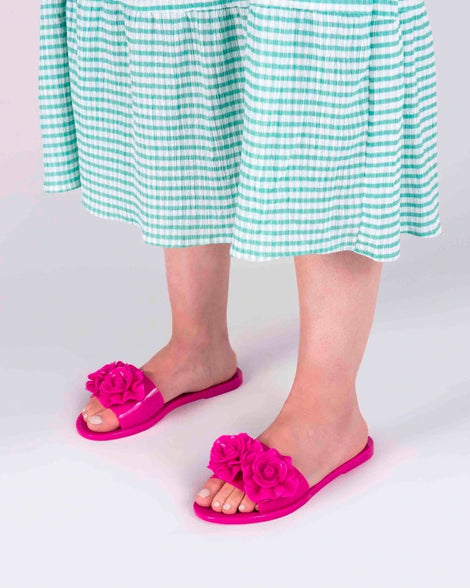 A model's legs wearing green gingham printed dress and a pair of neon pink Melissa Babe Garden slides with two flowers on the front strap.