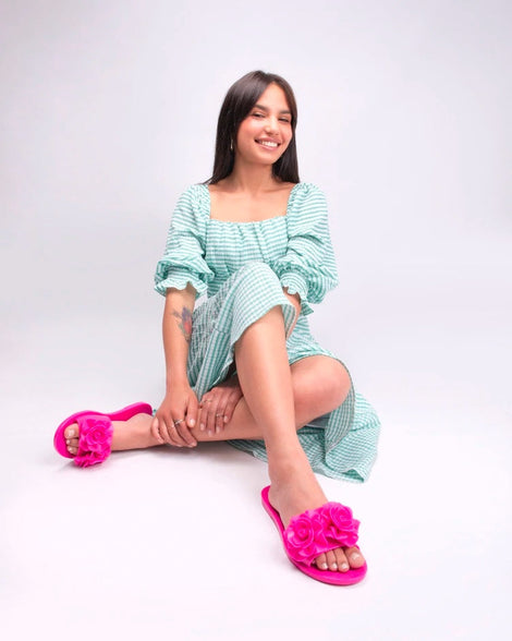 A model sitting on the ground with her legs crossed, wearing a greeen gingham printed dress, wearing a pair of neon pink Melissa Babe Garden slides with two flowers on the front strap.