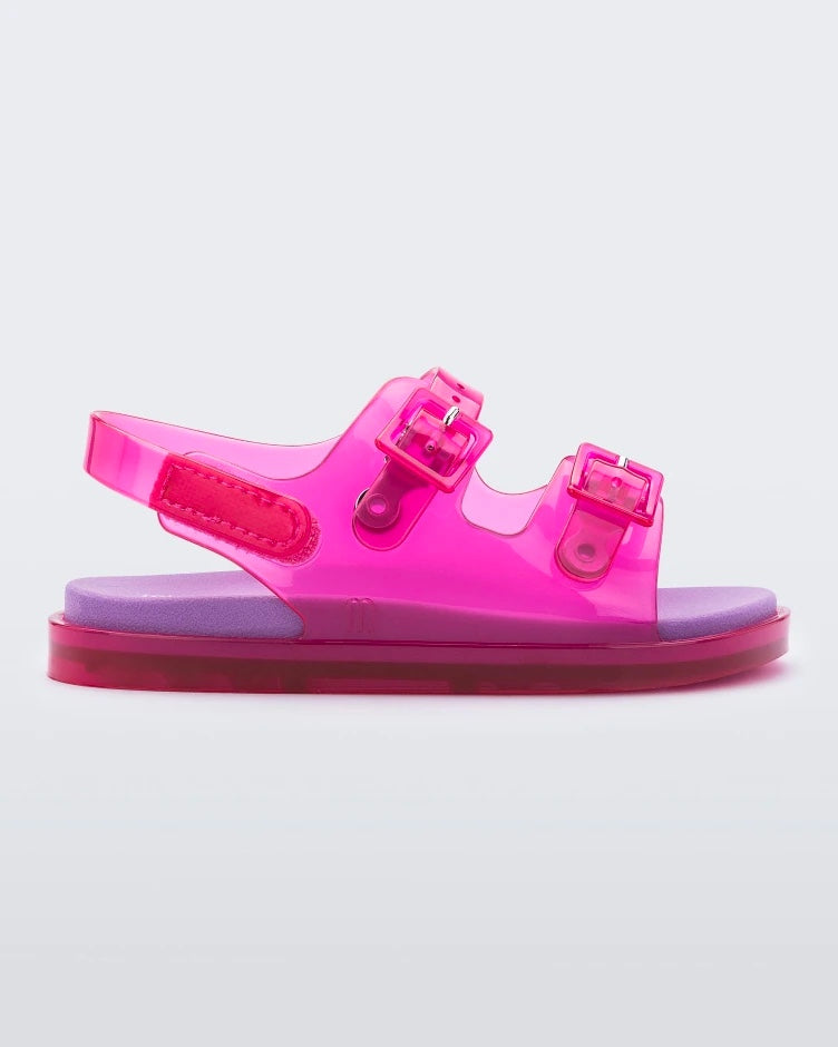 An outter side view of a pink/lilac Mini Melissa Wide Sandal with a pink base, two pink buckles on top, a back ankle strap and a lilac insole.