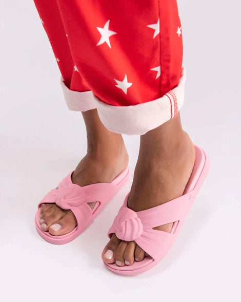 A model's legs in red patterned pants and a pair of pink Melissa Plush slides with a twist front detail.