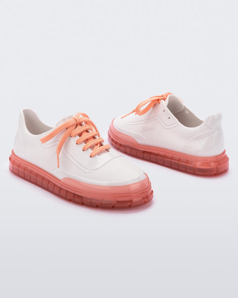 An angled side view of a pair of white Melissa Classic sneakers with a white base, laces, orange sole and a melissa logo on the side.