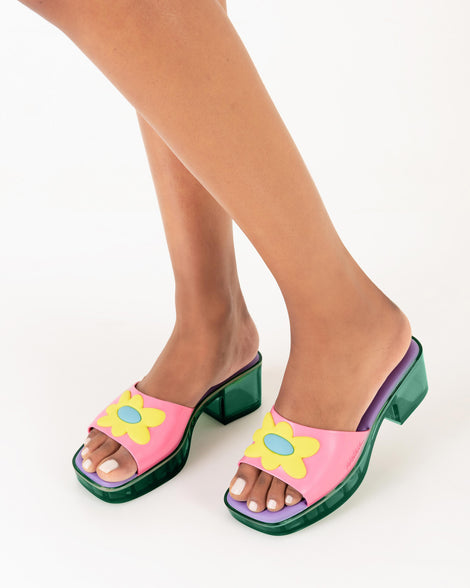 A model's legs wearing a pair of Melissa Shape slides with green platform heel,  a pink wide front strap with a yellow and blue flower on the top of the strap and a purple insole