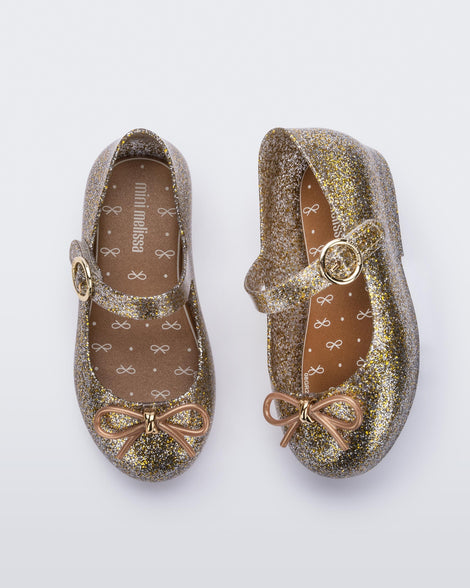 A top and side view of a pair of Gold/Silver/Glitter Mini Melissa Sweet Love flats with a top strap and a bow detail on the toe.