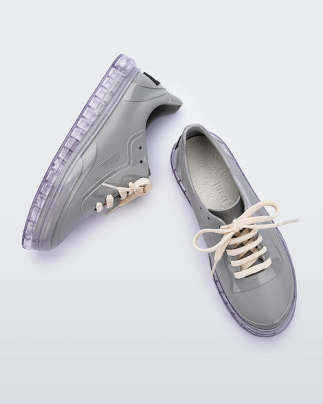 An angled top and side view of a pair of gray Melissa Classic sneakers with a gray base, beige laces, clear gray sole and a melissa logo on the side.