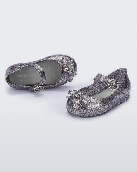 An angled front and top view of a pair of silver glitter Mini Melissa Sweet Love flats with a top strap and a bow detail on the toe.