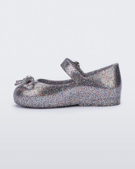 An inner side view of a silver glitter Mini Melissa Sweet Love flat with a top strap and a bow detail on the toe.