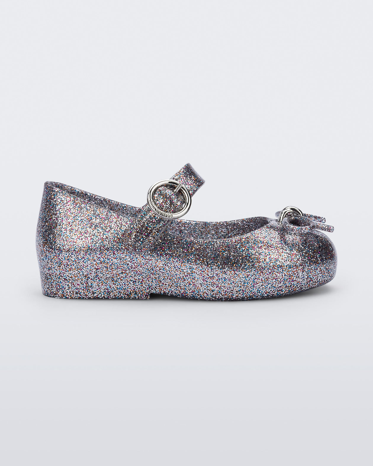 An outter side view of a silver glitter Mini Melissa Sweet Love flat with a top strap and a bow detail on the toe.