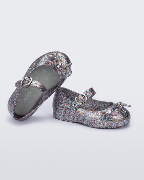 A side and top view of a pair of silver glitter Mini Melissa Sweet Love flats with a top strap and a bow detail on the toe, leaning on eachother