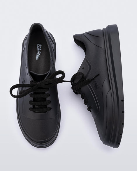 A top and side view of a pair of matte black Melissa Classic sneakers with a black base, laces, sole and a melissa logo on the side.