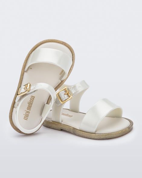 An angled top and side view of a pair of white Mini Melissa sandal with two straps and a gold buckle, leaning on eachother.