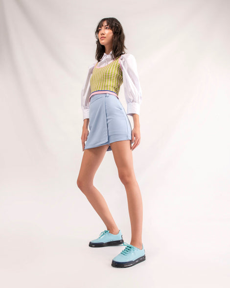 A model posing for a picture in a blue skirt, green and white shirt and wearing a pair of blue Melissa Classic sneakers with a blue base, laces and a black sole.