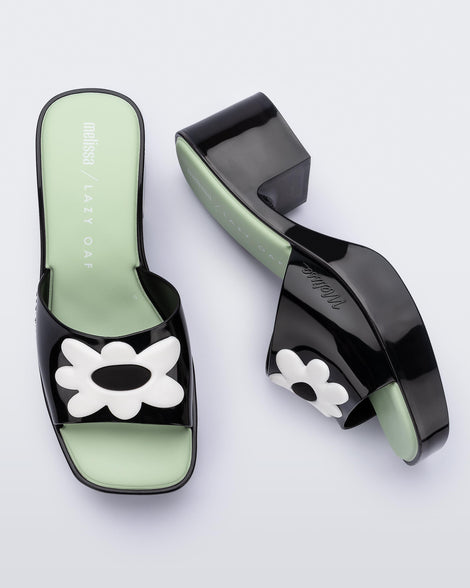 Top and side view of a pair of Melissa Shape slide sandals in black with platform heel, black front strap with white flower detail and a green insole. 