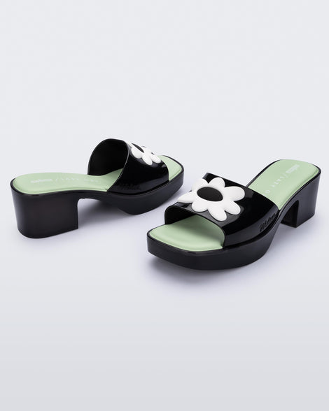 Angled view of a pair of Melissa Shape slide sandals in black with platform heel, black front strap with white flower detail and a green insole. 