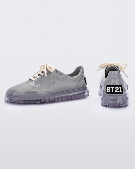 A side and back view of a pair of gray Melissa Classic sneakers with a gray base, beige laces, clear gray sole and a melissa logo on the side and a BT21 black and white logo on the back.