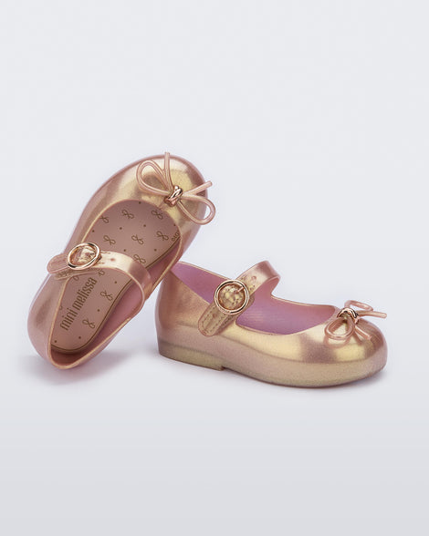 An angled side and top view of a pair of pink Mini Melissa Sweet Love flats with a top strap and a bow detail on the toe, leaning on one another.